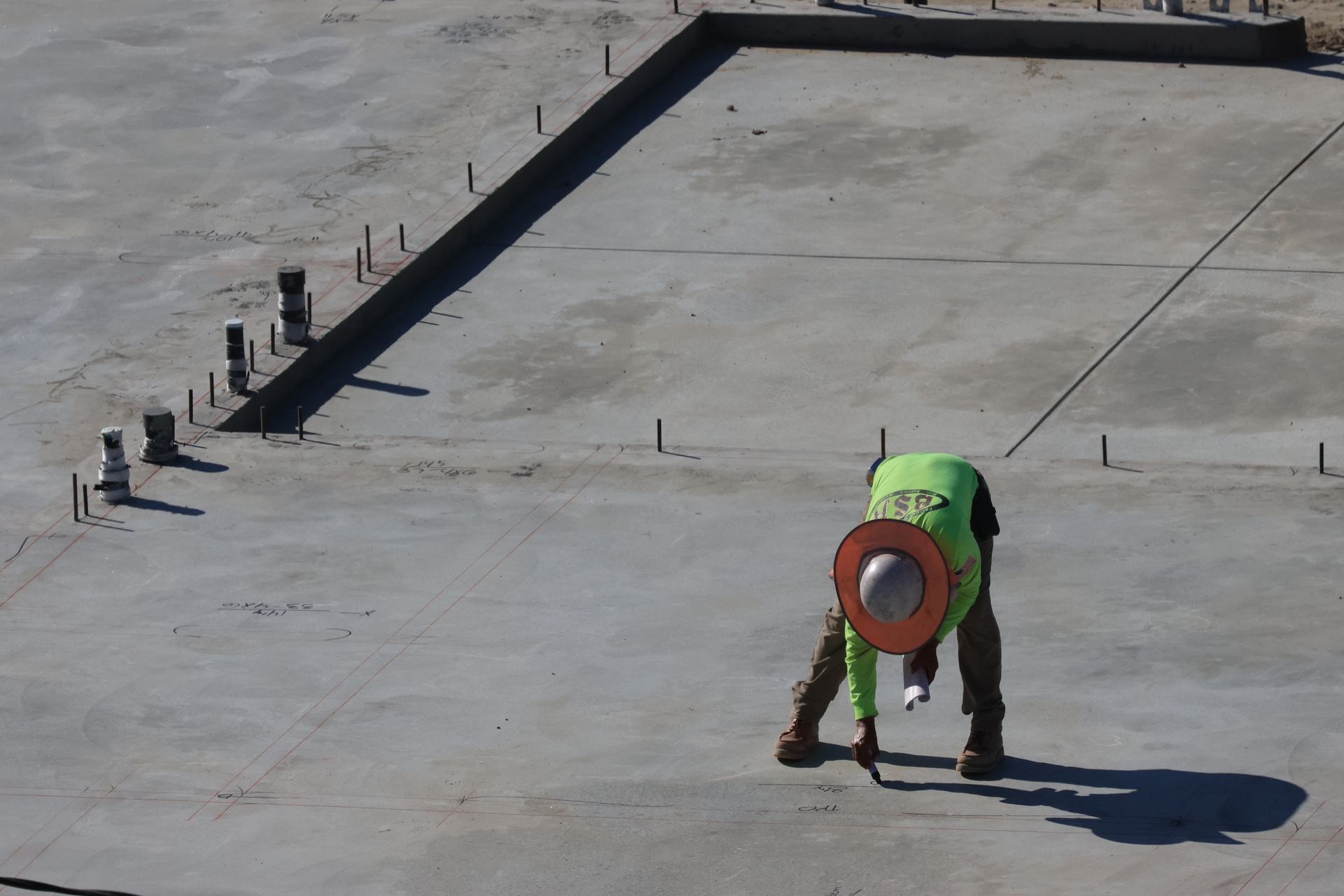 Worker marking layout lines across a large finished concrete slab with rebar dowels set along the edge