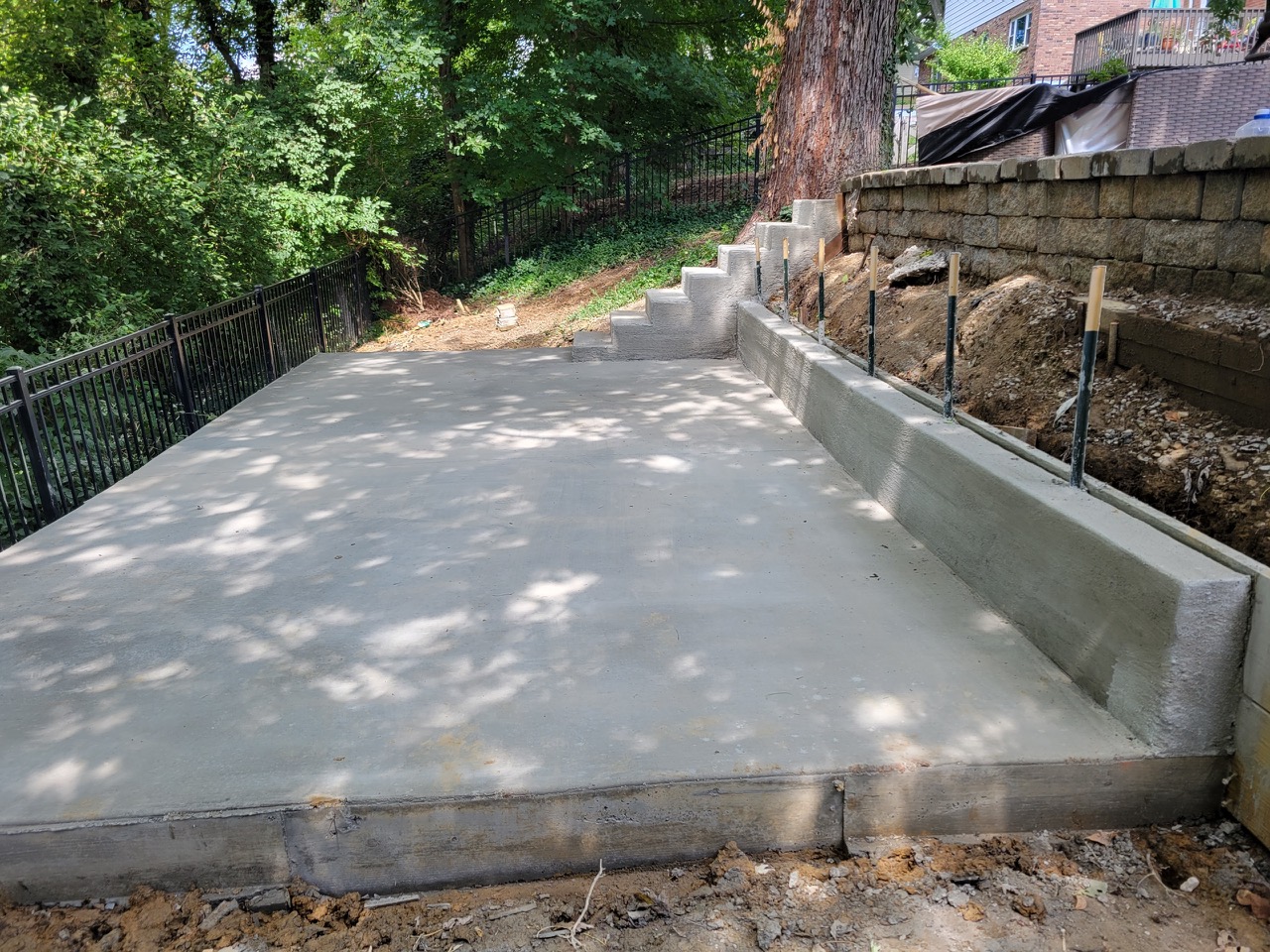 Concrete slab with a poured integral curb wall along one edge and concrete steps climbing the adjacent slope