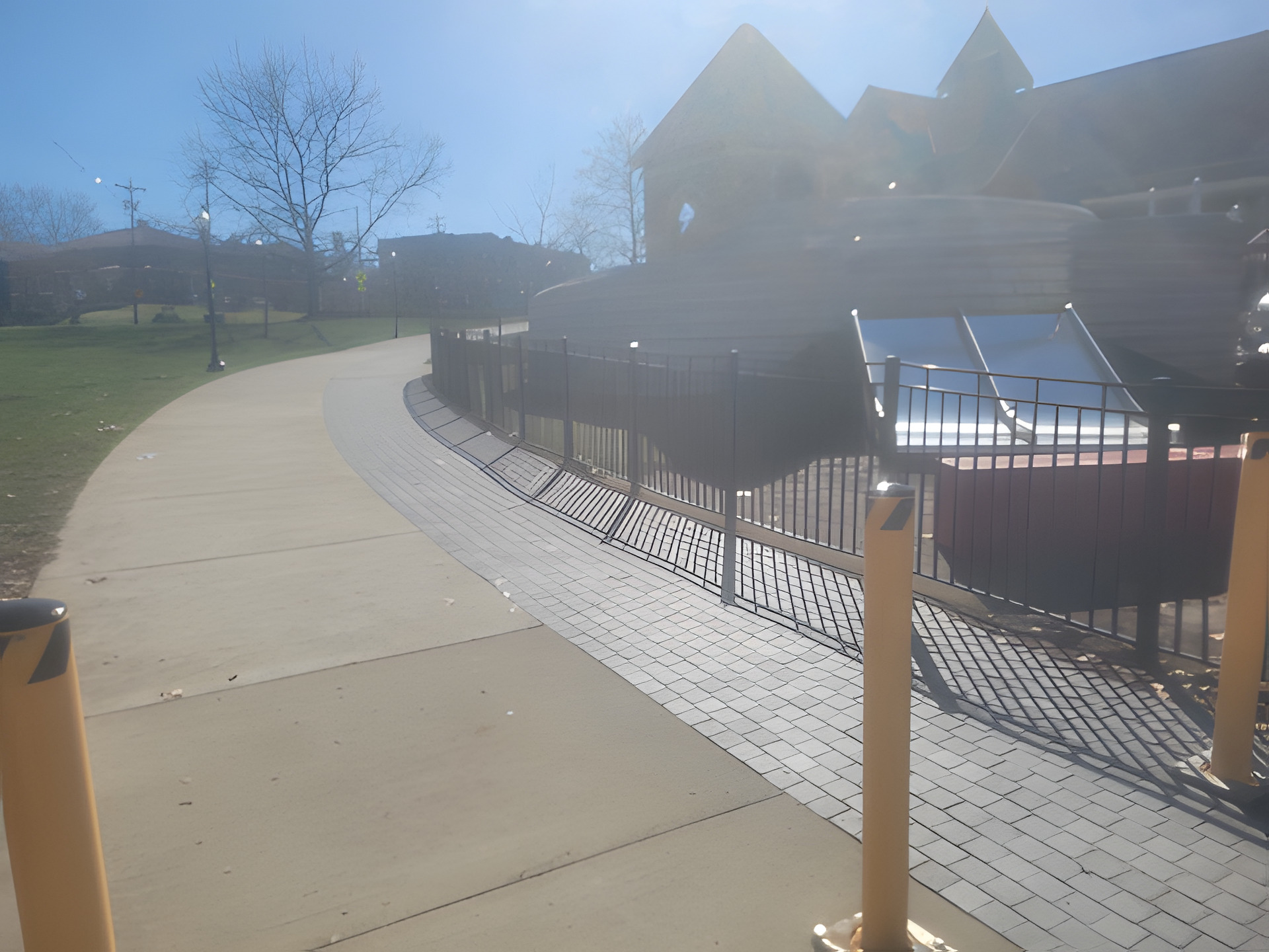 Concrete walkway curving through a public park alongside a paver band and protective bollards