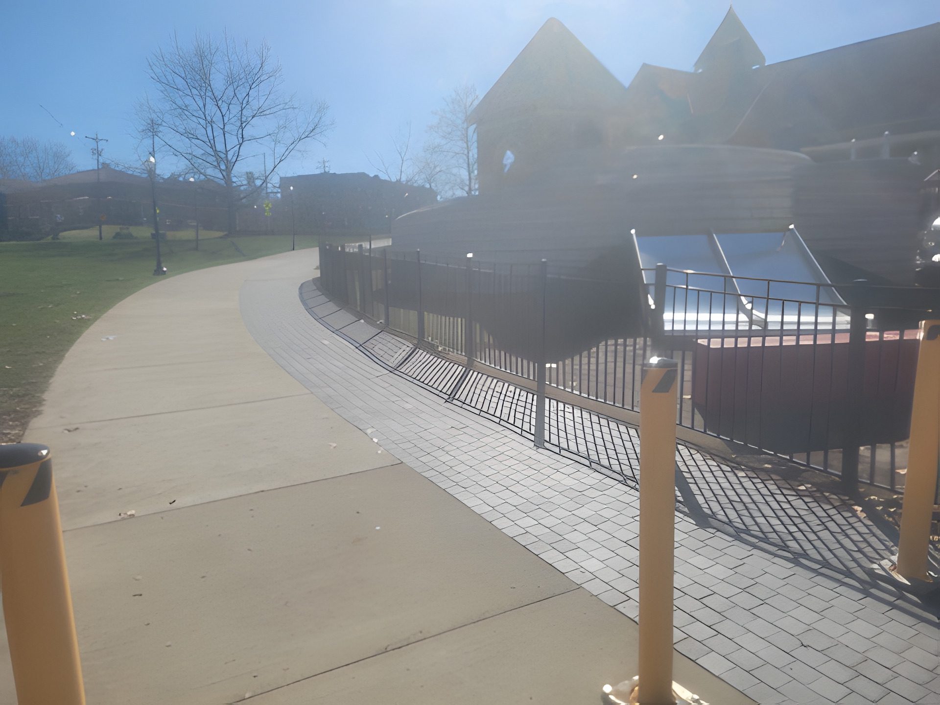 Concrete walkway curving through a public park alongside a paver band, protective bollards and fenced plaza